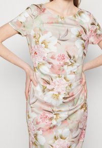 Kobieta nosząca dopasowaną sukienkę z krótkimi rękawami, zdobioną pastelowym kwiatowym wzorem w różowe i białe kwiaty z zielonymi i brązowymi liśćmi.