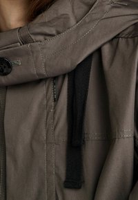 Zbliżenie na rękaw kurtki w kolorze taupe z czarnym guzikiem i czarnym sznurkiem przy ramieniu.