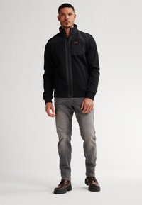 Chaqueta negra con cremallera y cuello alto, bolsillo frontal y paneles en contraste, combinada con pantalones vaqueros gris claro y botas marrones. Textura suave.