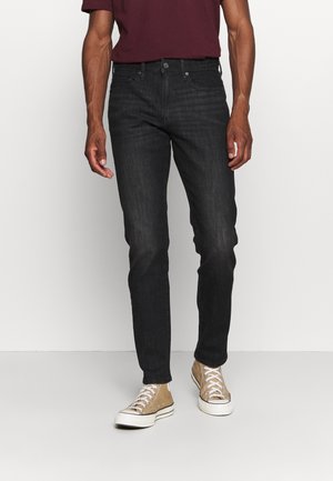 Man wearing slim-fit black jeans, beige high-top sneakers, and a maroon short-sleeve shirt standing against a white background.