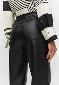 Czarne spodnie z sztucznej skóry o gładkiej fakturze, z dopasowanym pasem, dwiema tylnymi kieszeniami i luźnym krojem. Zdobione detalami w postaci przeszyć.