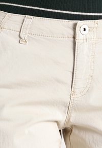 Ljusbeiga bomullsbukser med en slät textur, standardfickor och en metallknappknäppning i midjan.