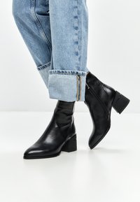 Bottines en cuir noir avec un bout pointu et un talon block, associées à un jean en denim bleu clair retroussé présentant une fermeture éclair contrastante.