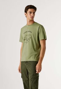 Camiseta de algodón verde oliva con un estampado gráfico negro de una furgoneta y texto, con cuello redondo y mangas cortas, combinada con pantalones verde oscuro.