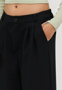 Black tailored trousers with a slight sheen, featuring a button closure, pleats, and an elastic waistband for comfort. Smooth texture.
