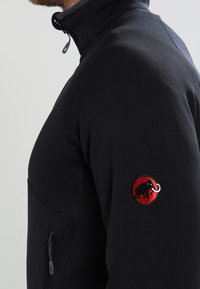 Man i en svart zip-up jacka med en röd cirkulär logo som föreställer ett mammut på överarmen mot en neutral bakgrund.
