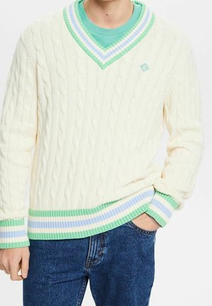 Person wearing a white cable-knit V-neck sweater with green, blue, and white striped trim, paired with blue jeans, hand in pocket.