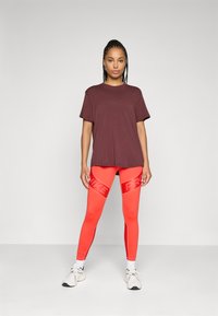 T-shirt corto color bordeaux abbinato a leggings arancioni fluorescenti con il marchio riflettente "Nike", completato da scarpe sportive bianche.
