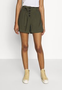Donna che indossa pantaloni corti verde oliva a vita alta con bottoni frontali e cintura con nodo, abbinati a sneakers beige alte e una maglietta bianca.