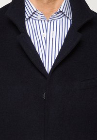 Granatowy płaszcz z wełny z domieszką, z zapięciem na jeden guzik, klapami i kieszeniami po bokach, noszony na białej koszuli w niebieskie paski.