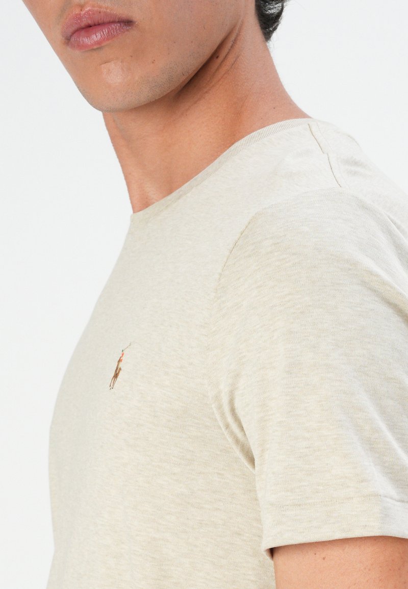 Camiseta de manga corta beige con un acabado texturizado y un pequeño logo bordado en el lado izquierdo del pecho. Presenta un diseño de cuello redondo.
