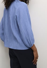 Blouse en tissu bleu clair froissé avec manches ballon et détail de bouton au poignet, associée à un pantalon foncé, sur un fond uni.