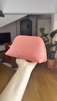 Rózsaszín bőrből készült kozmetikai táska texturált felülettel, lekerekített tetővel és cipzáras záródással. A logó látható az elején. Egy kéz tartja a táskát.