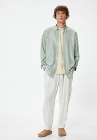 Camisa de botones a rayas verde claro, camiseta beige, pantalones de lino ligeros y zapatos de lona de color tostado; ajuste casual y textura de tela relajada.