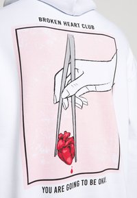 White hoodie featuring a graphic illustration of a hand holding a red heart with chopsticks against a pink background. Text below reads: "YOU ARE GOING TO BE OKAY."