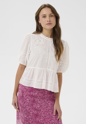 Vrouw met lang bruin haar die een witte geborduurde blouse met opgeblazen mouwen en een roze roks met patroon draagt, tegen een effen achtergrond.