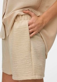 Beżowe, strukturalne szorty z elastycznym pasem i bocznymi kieszeniami, w połączeniu z luźną, dopasowaną koszulą na guziki. Lekka tkanina.
