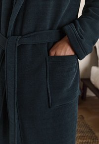 Tmavohnedý rebrovaný župan s viazaním v páse, ktorý má bočnú kapsu a mäkkú textúru. Detailný záber, ktorý zdôrazňuje textúru látky.