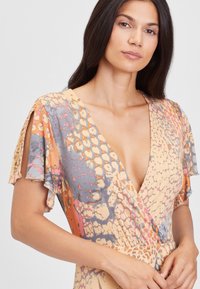 Sukňa s výstrihom do V, krátkymi rukávmi v žltej farbe s viacfarebným vzorom hada. Mäkká, ľahká látka s volánovými detailmi na rukávoch.