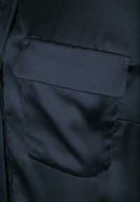 Tmavá saténová látka s hladkým povrchem; viditelná je jedna náprsní kapsa s klopou, která se integruje do celkového designu.