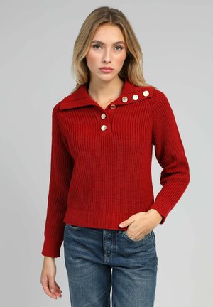 Pull rouge en tricot avec une texture côtelée, doté d'un col et de trois grands boutons sur une épaule. Associé à un jean en denim bleu.