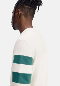 Suéter blanco con franjas horizontales verdes en las mangas. La tela parece suave y con textura, con un escote redondo y un diseño ajustado.