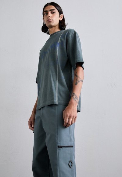 Camiseta de gran tamaño en color teal con mangas cortas y detalles en la costura lateral, combinada con pantalones grises ajustados que cuentan con un bolsillo con cremallera y un acento en forma de rombo.