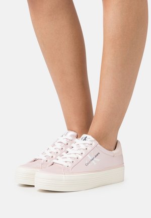 Baskets rose clair en matériau synthétique lisse, avec des lacets blancs, un logo sur le côté et une semelle blanche épaisse et texturée.