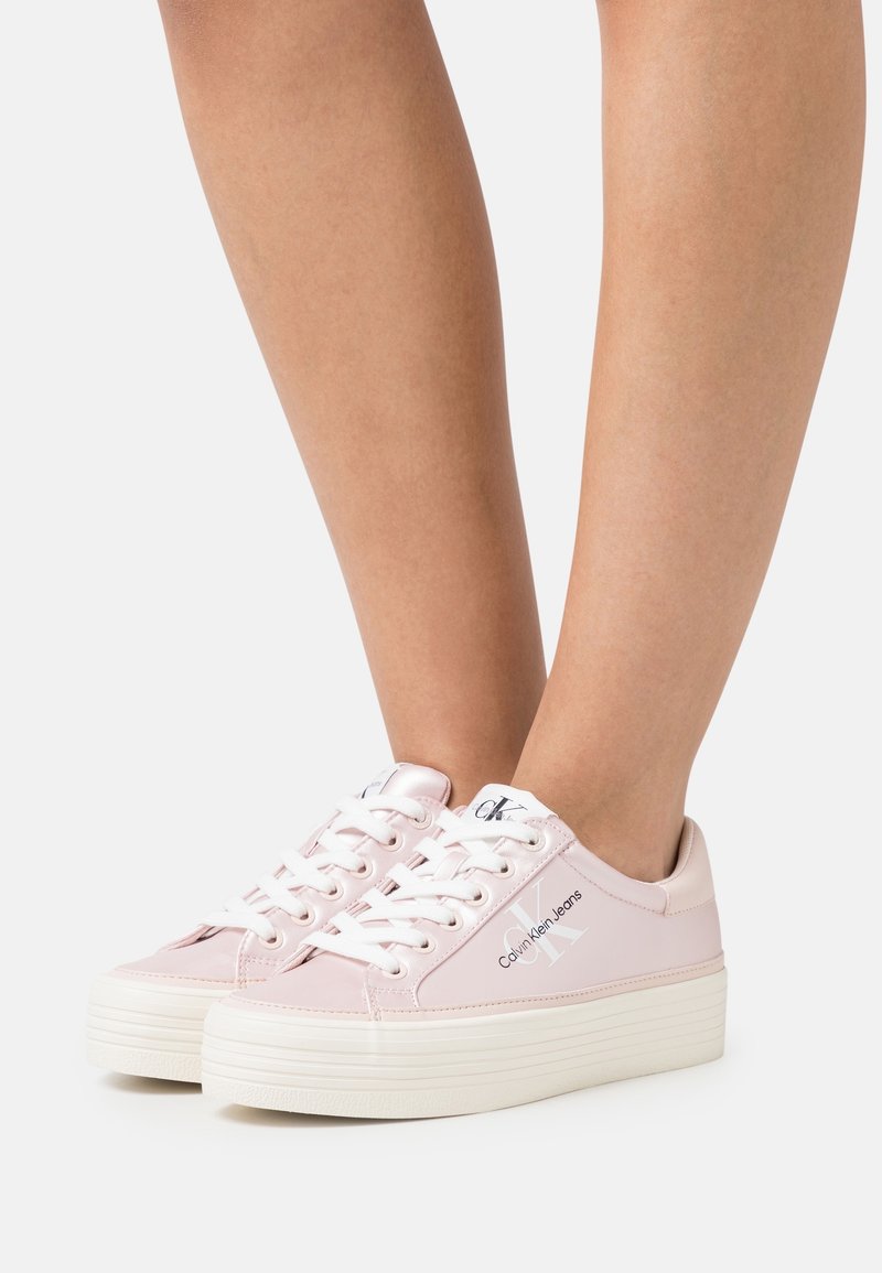 Baskets rose clair en matériau synthétique lisse, avec des lacets blancs, un logo sur le côté et une semelle blanche épaisse et texturée.
