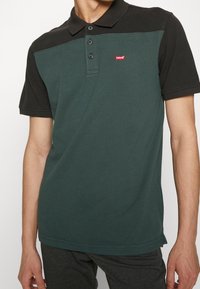 Polo tričko s tmavě zeleným tělem, černými krátkými rukávy, třemi knoflíky na léze a malým červeným logem Levi's na prsou.