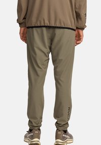 Persoon in olijfgroene joggpantalon en bijpassende jack, staand met handen langs de zij, zichtbaar RVCA-logo op het broekbeen.