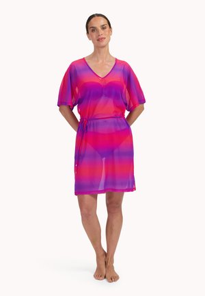 Vrouw die blootsvoets staat, gekleed in een korte, doorschijnende jurk met roze en paarse strepen, een taille met trekkoord en een V-hals.