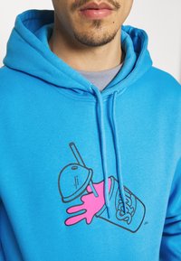 Turkos hoodie med en grafik av en kopp med en rosa hand och sugrör. Slät textur med dragsnoddar och en avslappnad passform.