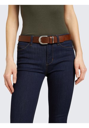 Henkilö pukeutuneena tummansinisiin farkkuihin, ruskeaan nahkavyöhön ja oliivinvihreään istuvaan toppiin, kädet rentoina sivuilla.