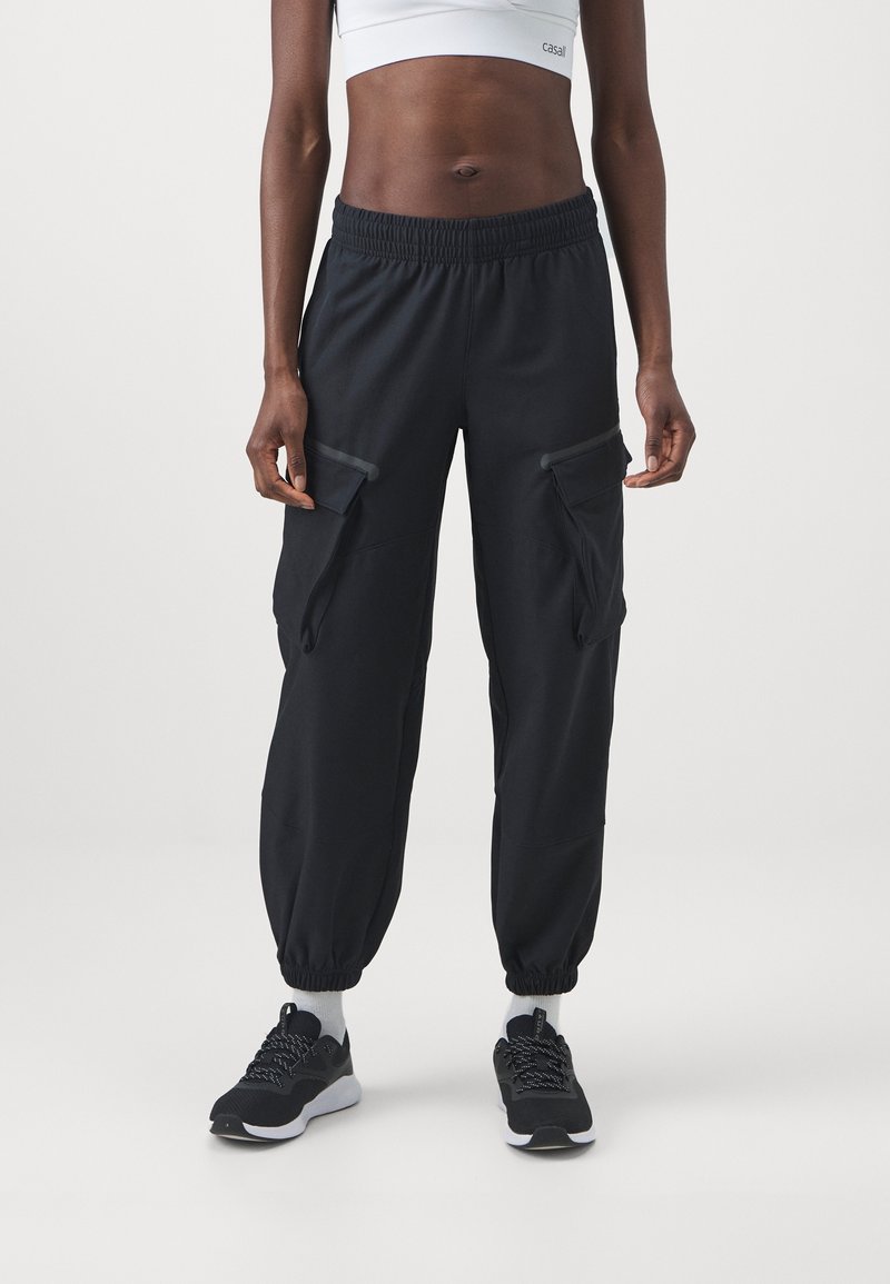 Personne portant un pantalon cargo noir à taille élastique avec des poches latérales, des chaussettes blanches, des baskets noires et un top de sport court blanc, debout devant un fond clair.