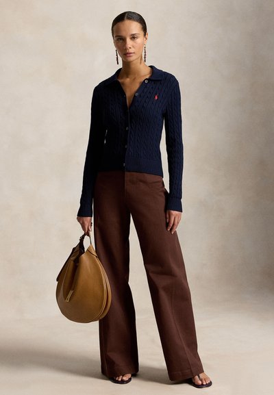 Nő tengerészkék, kötött mintás kardigánban, barna bőszárú nadrágban, szandálban, és egy nagy barna bőr táskát tart a kezében.