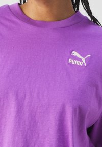 Fialové bavlněné tričko s kulatým výstřihem a malým bílým logem Puma na hrudi. Měkká textura a volný střih.