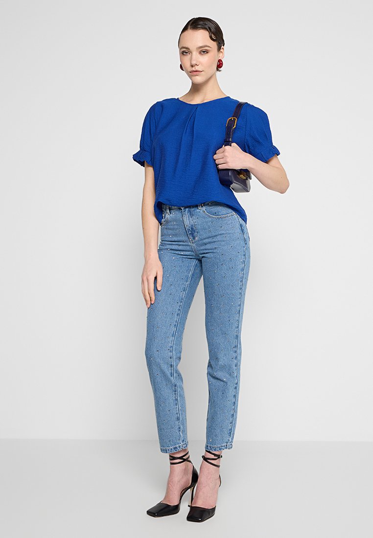 Top corto azul con mangas abullonadas, combinado con jeans azul claro que tienen sutiles adornos. Llevado con tacones negros de tiras.