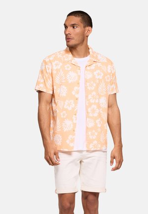 Hombre de pie mirando hacia la izquierda, con una camisa de manga corta color melocotón con estampado floral sobre una camiseta blanca y pantalones cortos blancos, frente a un fondo liso y claro.