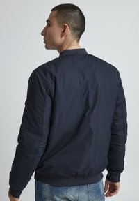 Chaqueta bomber azul marino con un tejido suave y ligero, cuello y puños acanalados, y un bolsillo con cremallera en la manga izquierda.