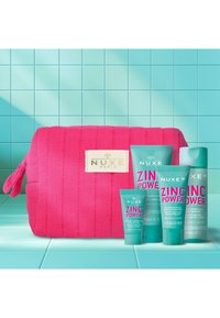 Neceser de cosméticos acolchado en color rosa con etiqueta de NUXE, que contiene cuatro productos de cuidado de la piel de zinc en color turquesa con texto en negrita rosa, colocado sobre una superficie de baldosas azul claro.