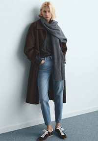 Cappotto lungo marrone, sciarpa grigia, maglione grigio, jeans azzurri e sneakers scure con lacci bianchi. Materiali lavorati e vestibilità comoda.