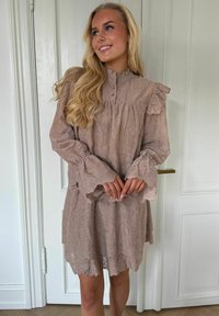 Tânără cu păr lung și blond poartă o rochie taupe din dantelă, cu mâneci și guler cu volane, stând în fața unor uși albe cu panouri.