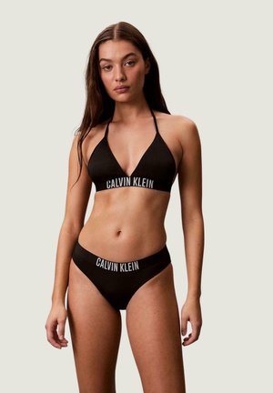 Joven mujer vestida con bikini negro de Calvin Klein con logo en la parte superior e inferior, de pie frente a un fondo claro y liso.
