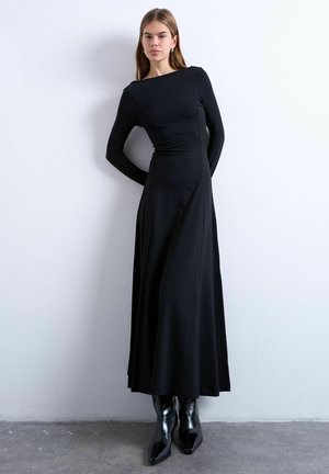 Vrouw staat tegen een muur, gekleed in een lange zwarte nauwsluitende jurk met lange mouwen en zwarte glanzende puntige laarzen.