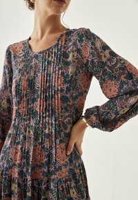 Vestido con estampado floral que presenta pliegues, mangas largas y un escote redondo. Los colores incluyen verde oscuro, rosa y naranja sobre un fondo oscuro.