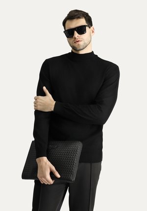 Zwarte gebreide trui met hoge hals, gedragen met zwarte broek. Houdt een gestructureerde zwarte clutch met een geweven patroon vast.