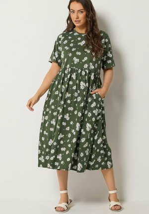 Kvinde iført en grøn midikjole med hvide tusindfrydtryk, korte ærmer, sidelomme og hvide ankelremssandaler, stående op ad en hvid væg.