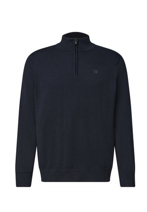 Homme aux cheveux courts, bouclés et foncés, portant un pull bleu marine à fermeture éclair quart sur une chemise blanche et un jean bleu, debout devant un fond uni.
