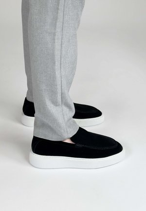 Persoon die lichtgrijze broek en zwarte instapschoenen met dikke witte zolen draagt, staand op een effen lichte achtergrond.